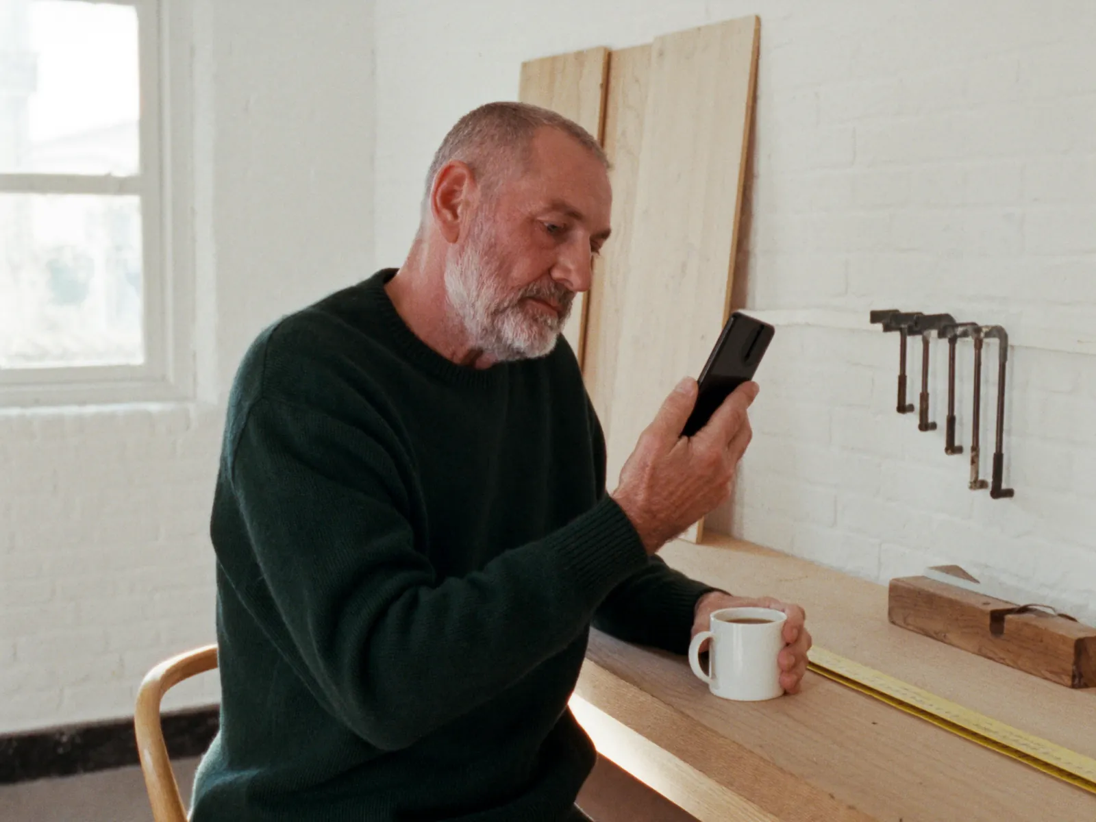 Illustration: tømrer holder kaffepause ved arbejdsbænken i sit værksted og læser på telefonen